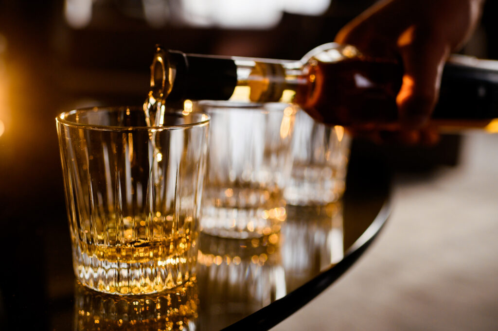 Close up of anonymous man pouring gold whiskey from bottle in glass which standing on wooden table indoors